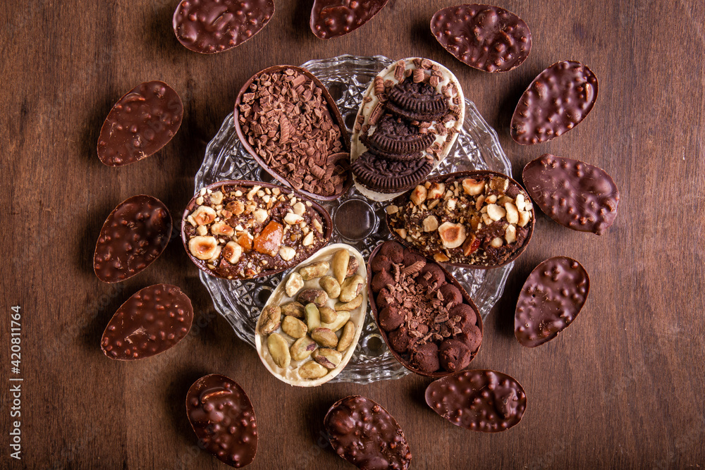 Fototapeta premium Top view of six mini stuffed easter eggs on a glass plate and mini crunchy stuffed chocolate with peanut easter eggs around it on a wooden table.