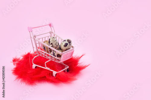 Beautiful Easter composition with shopping cart, quail eggs and red feathers against pink background with copy space. Easter sale concept. Selective focus photo