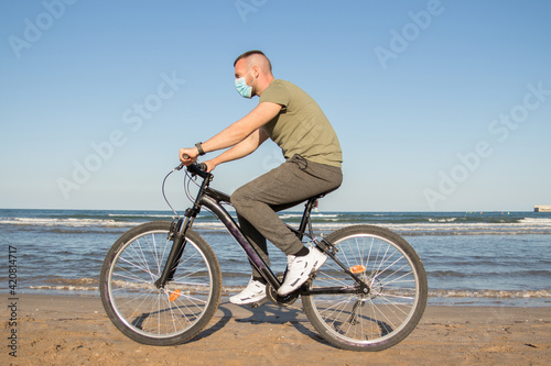 Boy with green t-shirt and mask walking along the beach on his bike