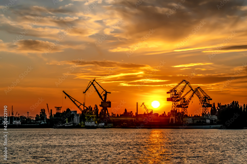 Sunset silhouette of Kherson port (Ukraine). The sun sets behind the port cranes against the background of an orange sky and reflections in the water of the Dnieper River