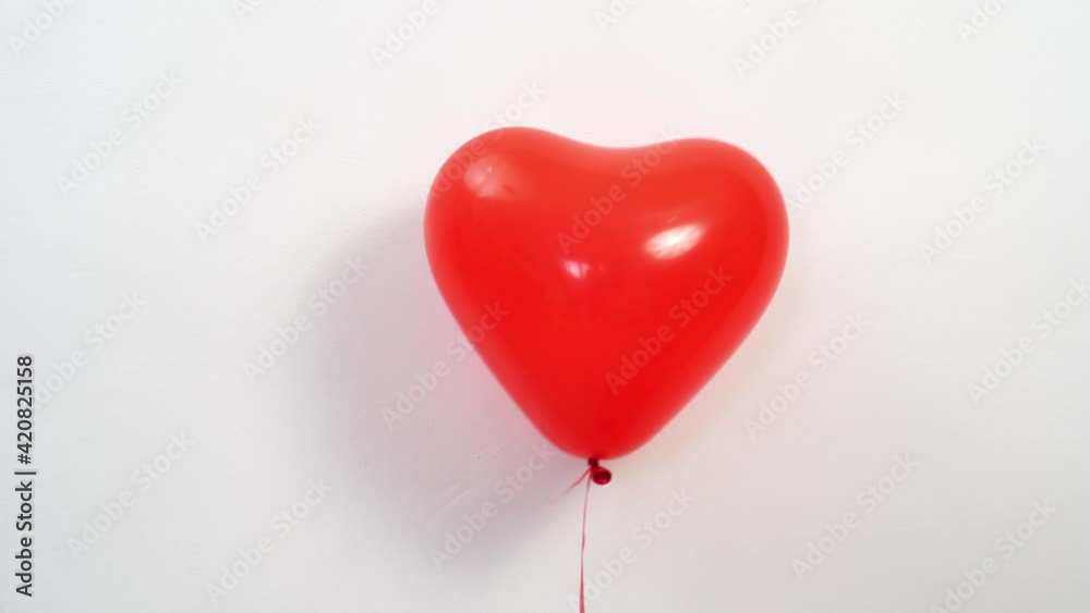 A heart shaped red balloon on a rope against a white wall flies off up. A heart shaped red balloon on a rope against a white wall flies off up.
