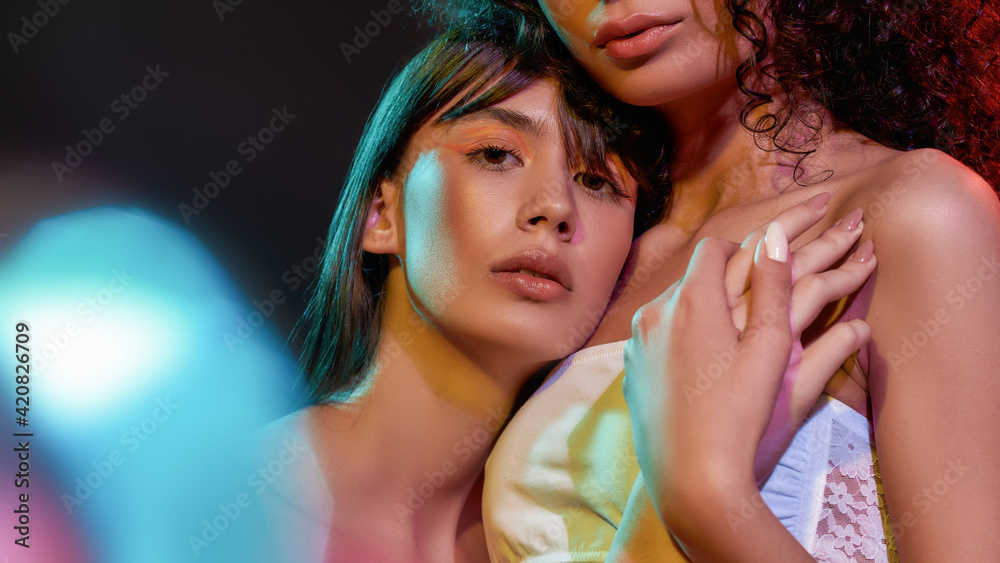 Close up portrait of two artistic young female models with professional art makeup hugging, posing together in neon light isolated over black background