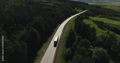 One Semi Truck with white trailer and cab driving, traveling alone on dense flat forest asphalt straight road, highway top view follow vehicle aerial footage. Freeway trucks traffic