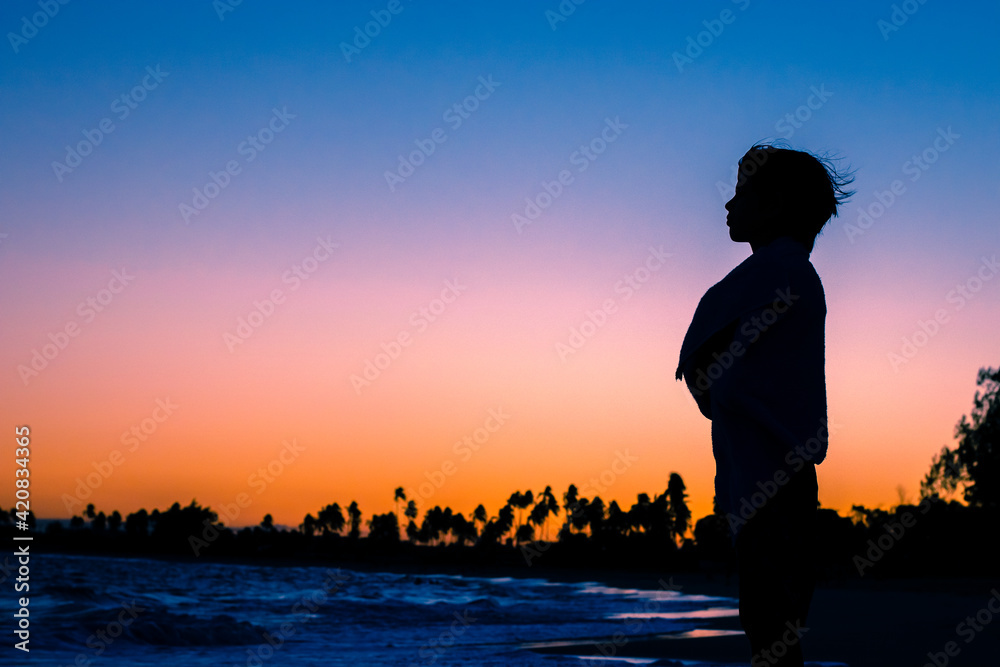 Silhueta de uma criança enrolada numa toalha olhando para o horizonte durante lusco fusco do pôr do sol em uma praia.