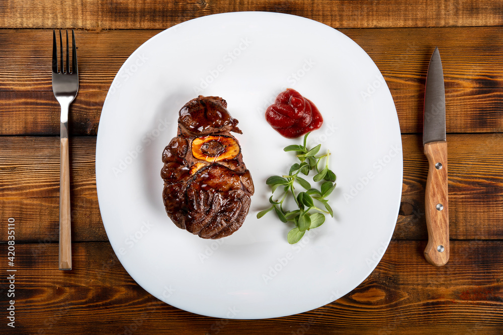 Grill the meat with a bone on a white plate. Sauce, herbs. Knife, fork. Wooden table. Grill the meat with a bone on a white plate. Sauce, herbs. Knife, fork. Wooden table.