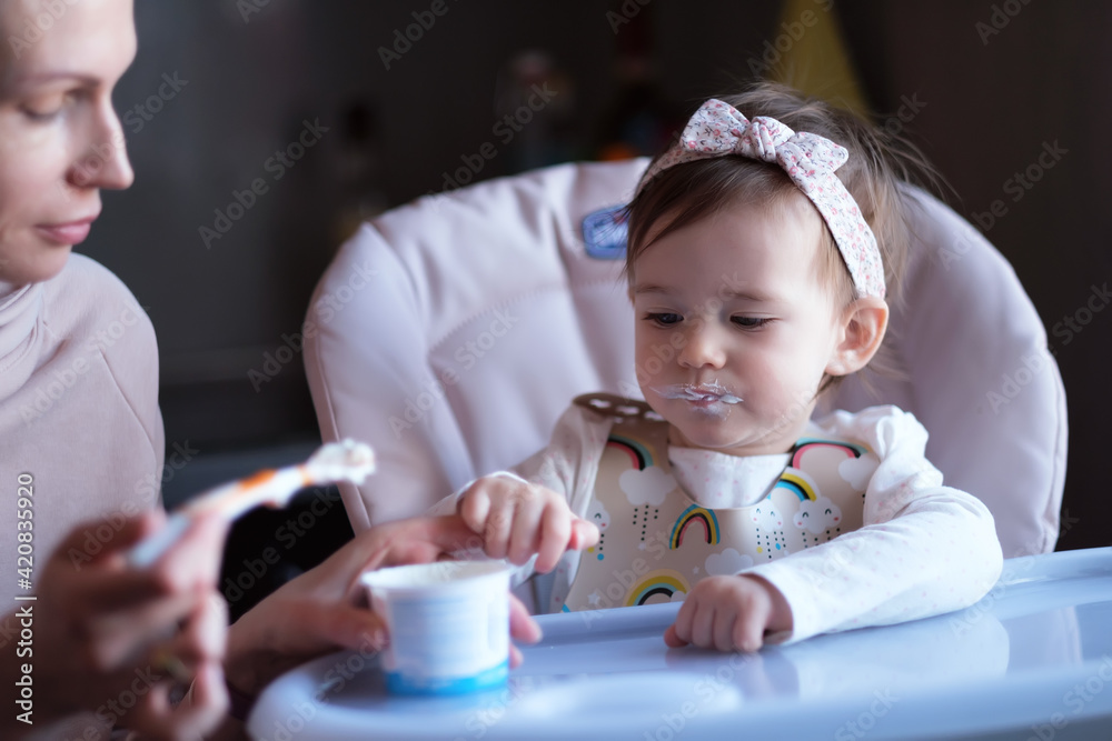 Mom was spoon-feeding the baby girl in the highchair. Girl looking at the camera.Family lifestyle shooting. Selective focus. Mom was spoon-feeding the baby girl in the highchair. Girl looking at the camera.Family lifestyle shooting. Selective focus.