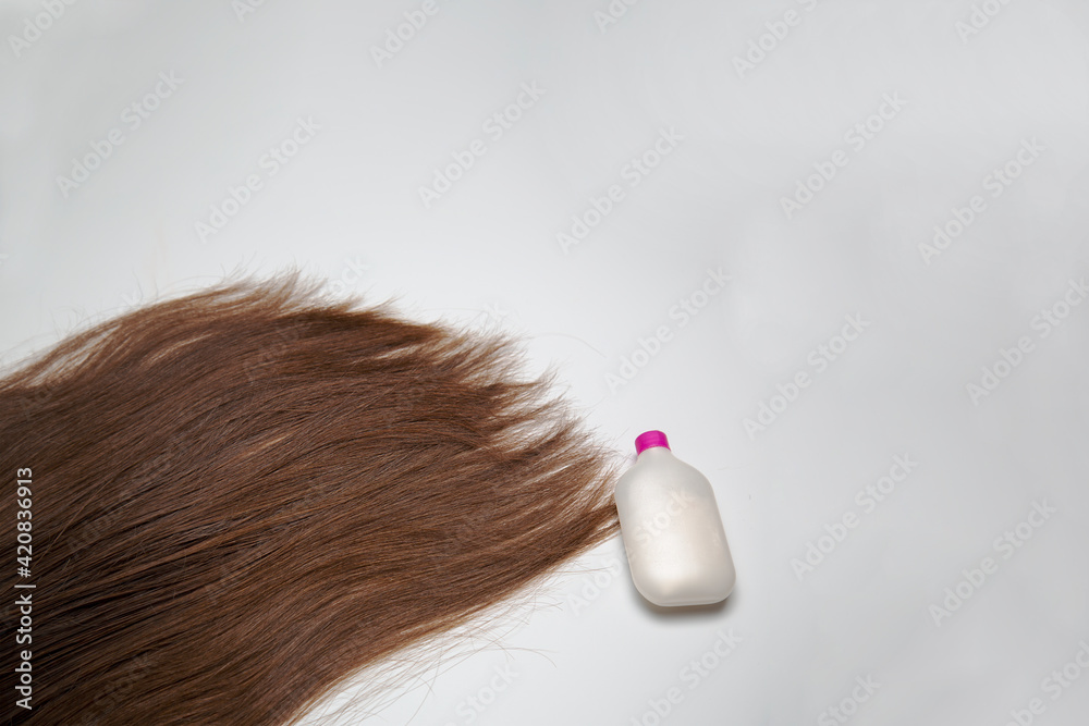 Thick, long, natural hair lies on a white surface. Brown hair on a white background. Top view. A white tube of shampoos, a bottle of balsam. Hair care and treatment. Thick, long, natural hair lies on a white surface. Brown hair on a white background. Top view. A white tube of shampoos, a bottle of balsam. Hair care and treatment.