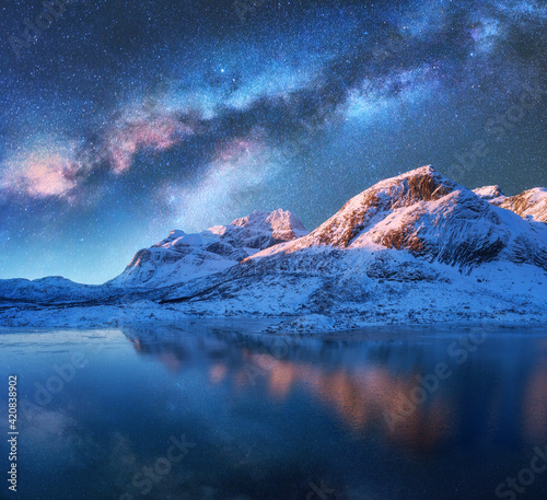 Milky Way above frozen sea coast and snow covered mountains in winter at night in Lofoten Islands, Norway. Arctic landscape with blue starry sky reflected in water, snowy rocks, milky way. Space