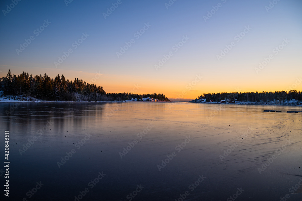 A peaceful winter scenery of frozen seashore with a hut in a chilly weather with clear sky horizon, sunrays and pine trees