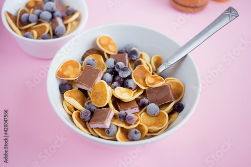 Pancake cereal in a plate with blueberries and chocolate on a pink table.Trendy modern breakfast with little pancakes.
