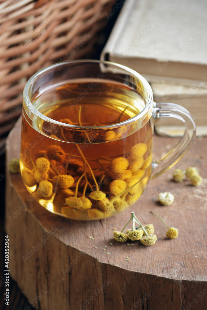 Tansy seed tea or infusion in a glass cup with flowers on wood with books nearby, herbal drink is good as anthelmintic, menstrual, kidney, rheumatism remedy , closeup, naturopathy, homeopathy concept