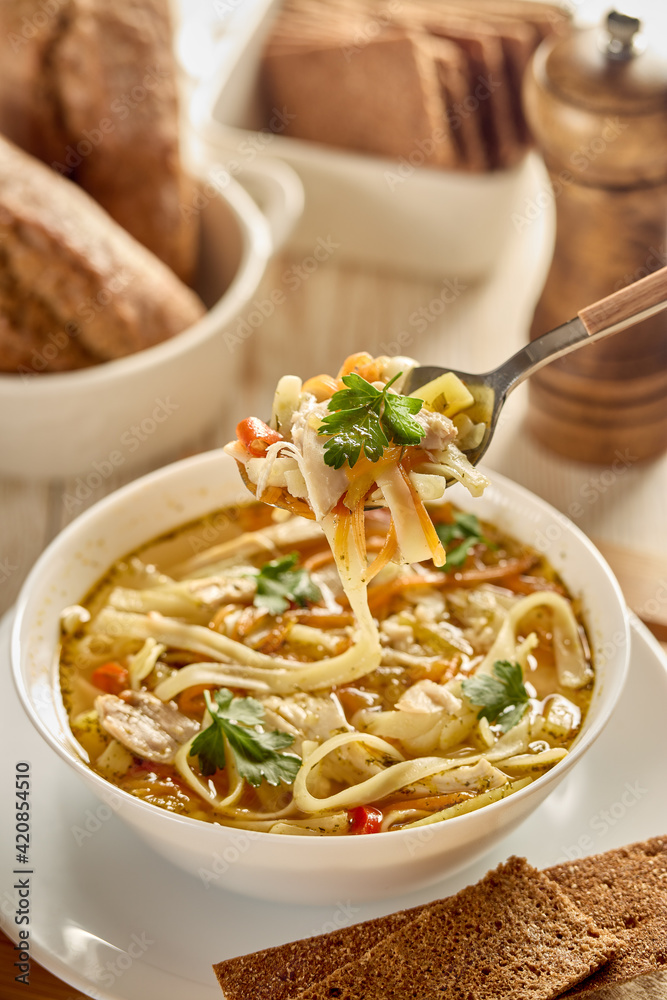 Soup with noodle, pieces of chicken and vegetables in white bowl served with spices and breads on light wooden background. Soup with noodle, pieces of chicken and vegetables in white bowl served with spices and breads on light wooden background.