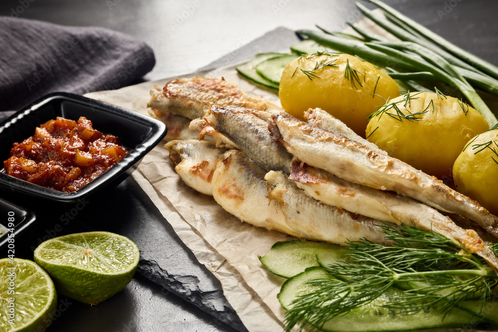 Fried smelts serving with potatoes, vegetables, lime and sauces. Grey slate background. Selective focus on fish. Fried smelts serving with potatoes, vegetables, lime and sauces. Grey slate background. Selective focus on fish.