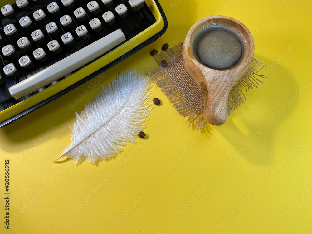 on a yellow background there is a typewriter Jerzy with an ostrich feather. Next to it is a wooden cup with coffee and scattered coffee beans - obrazy, fototapety, plakaty 