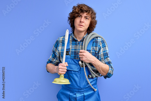 confident plumber man with curly hair in uniform holding toilet plunger isolated on blue background © alfa27