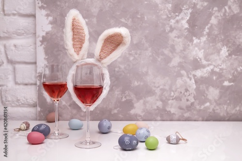 Two glasses of rose or red wine with bunny ears and Easter decorations, colorful eggs on white table, on bright background. Copy space for text.