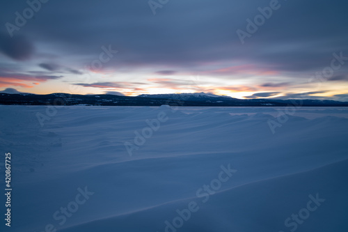 Winter frozen lake scene in northern Canada on a stunning cloudy sunset afternoon in March with white snow  mountains in background and iconic Canadian landscape in the north. 
