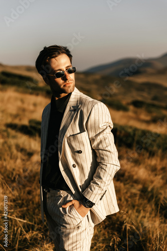 Handsome man in glasses and a light beige suit stands posing against a background of mountains
