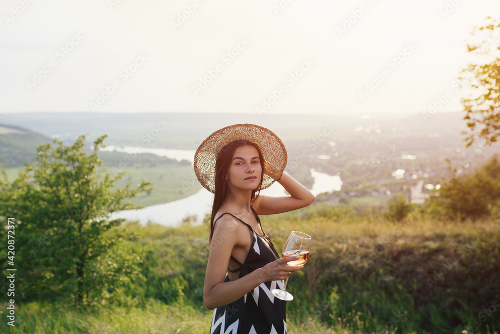 A beautiful attractive young woman in a elegant dress and straw hat drinks wine on the hill on a picnic. Summer vacation.