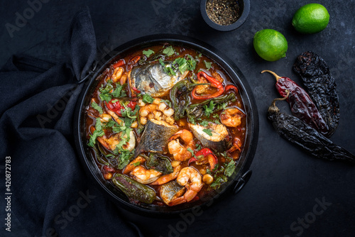 Modern style traditional Mexican seafood pozole soup with fish, king prawns and hominy in a clear sauce served as top view in design pot