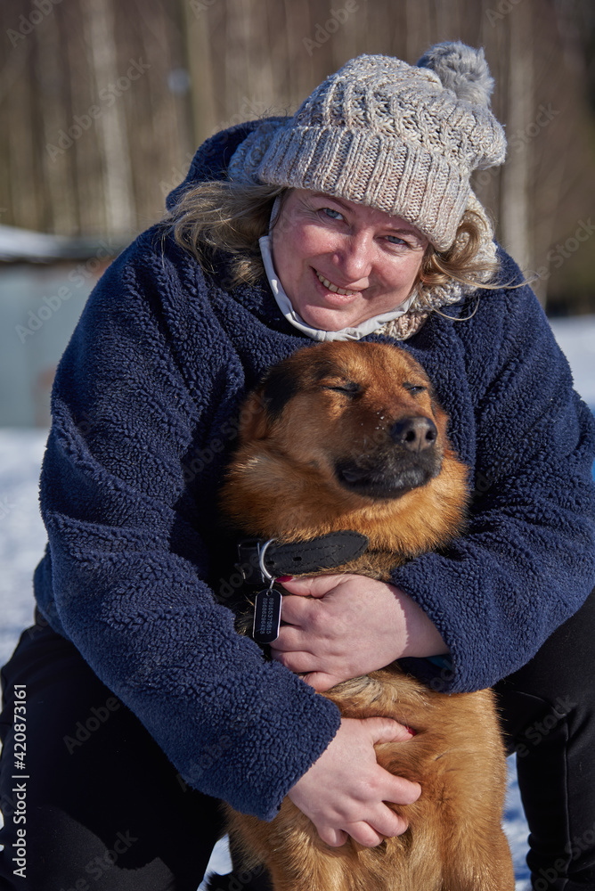 A plump middle-aged blonde woman in a knitted hat hugs a mongrel sitting on white snow on a sunny winter day.