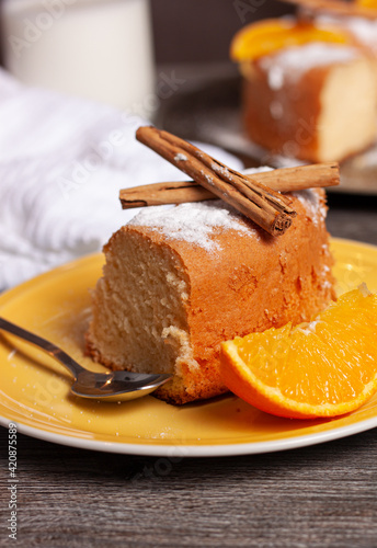 Concepto de un plato amarillo con un trozo de pastel con azúcar glash, ramas de canela, una rodaja de naranja y una cuchara de postre sobre un soporte de madera gris. photo