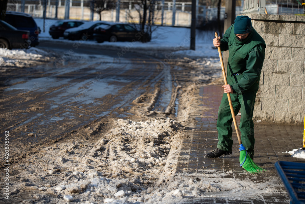 A manual worker sweeps the remnants of snow off a wet pavement with a broom A manual worker sweeps the remnants of snow off a wet pavement with a broom