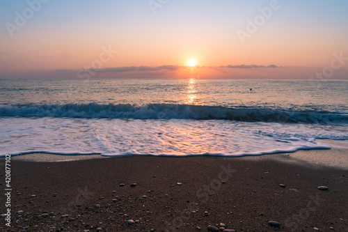 A beach in south Turkey at sunrise time with sand and pebbles in the foreground  two people as silhouettes in the background  motion blur of the waves