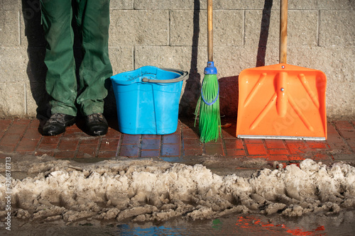A broom and a snow shovel lean against the wall and a blue bucket and a manual worker stand on the paving stone
