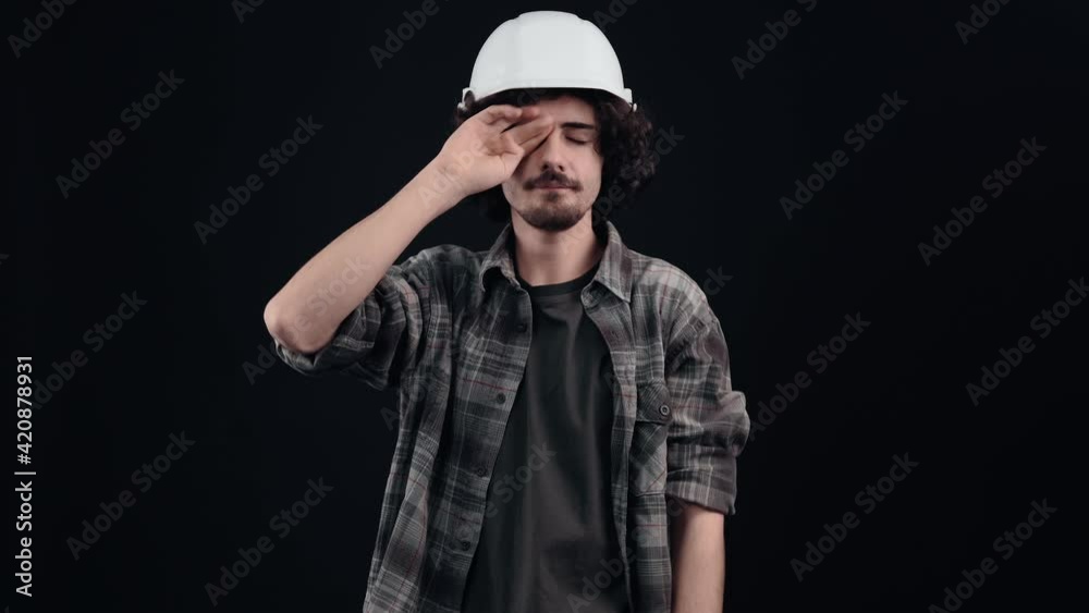 Sleepy hipster lies with his hands up then helmet looking into the room, bored. Young man with curly hair wears a special helmet on his head, dressed in a plaid shirt, on a black background, in the Sleepy hipster lies with his hands up then helmet looking into the room, bored. Young man with curly hair wears a special helmet on his head, dressed in a plaid shirt, on a black background, in the