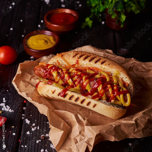 Very tasty traditional american fast food - hotdog. Grilled bun with sausage, mustard and ketchup, seasoning and tomatoes on dark background with copy space. 