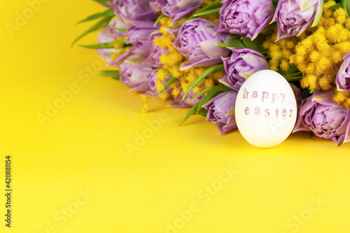 One white chicken egg with words Happy Easter written with help of stamps and pink metallic paint, bouquet of tulips and mimosas on yellow background, copy space. DIY, festive concept. Side view