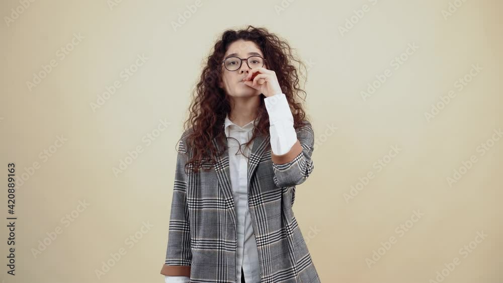 The young woman promises silence, keeps a secret in secret, like a basket with her hand on her lips. Cretaceous in gray jacket and white shirt, with glasses posing isolated on a beige background in