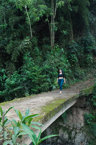 beautiful latin woman crossing a cement bridge covered with green moss in the middle of the Colombian jungle  observing nature feeling free and without fear of the void  height .
