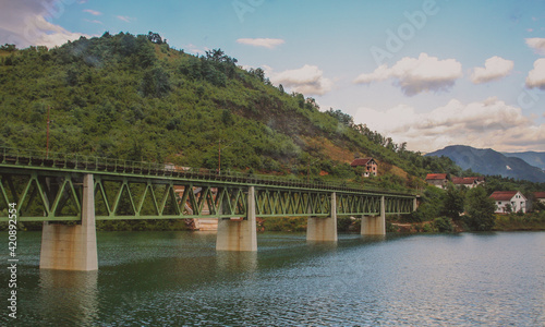 Colorful picture of a metal trestle bridge in Bosnia over a wide river. Five spans of bridge are visible.