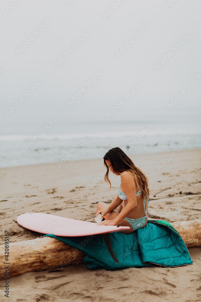 Young female surfer sitting on log waxing surfboard at beach, Ventura