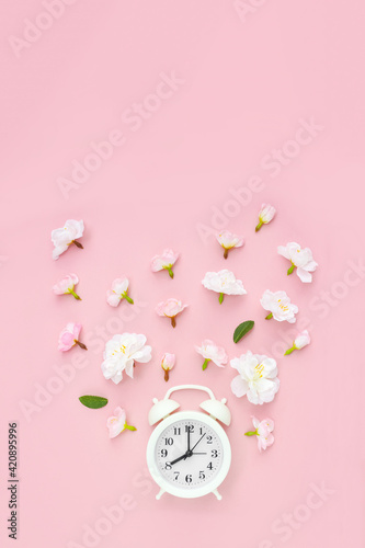 Alarm clock is decorated with gentle flowers on pink background. Summer time concept. Vertical poster. Copy space, layout, top view