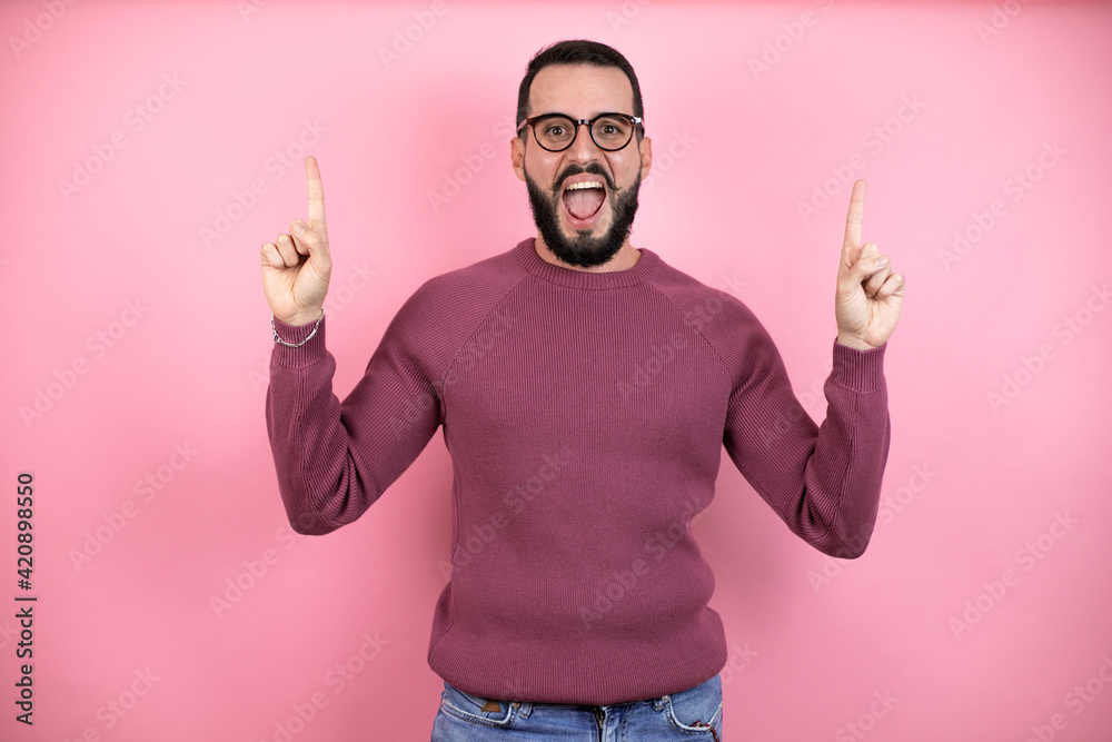 Handsome man wearing glasses and casual clothes over pink background amazed and surprised looking at the camera and pointing up with fingers and raised arms Handsome man wearing glasses and casual clothes over pink background amazed and surprised looking at the camera and pointing up with fingers and raised arms