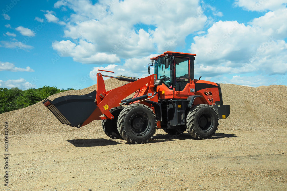 A red-black front-end loader with small wheels against the background of a large pile of stone sand and a blue sky with white clouds. Side view. A red-black front-end loader with small wheels against the background of a large pile of stone sand and a blue sky with white clouds. Side view.