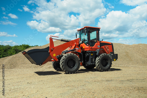 A red-black front-end loader with small wheels against the background of a large pile of stone sand and a blue sky with white clouds. Side view.