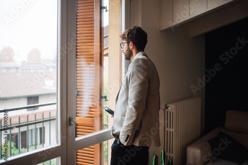 Mid adult man holding smartphone gazing out through apartment window photo