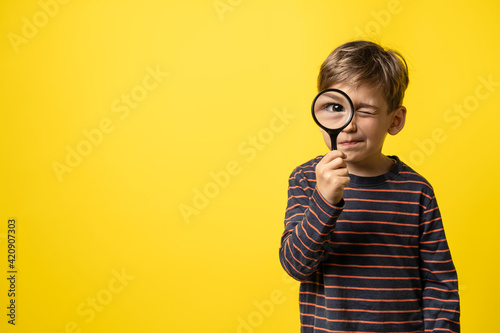 Front view portrait of small caucasian boy curious child holding a magnifying glass for reading in hand inspecting or investigating to have close look with eye in focus - copy space yellow background