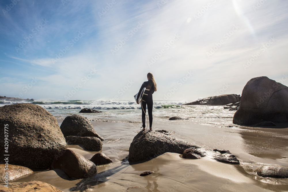 Young female surfer looking out from beach rock, rear view, Cape Town, Western Cape, South Africa Young female surfer looking out from beach rock, rear view, Cape Town, Western Cape, South Africa