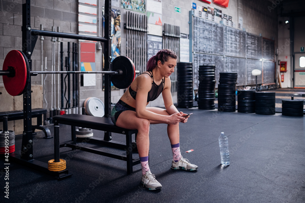 Young woman training, sitting on weights bench in gym looking at smartphone Young woman training, sitting on weights bench in gym looking at smartphone