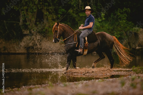 White man riding a horse inside a river, wearing a straw hat, with his horse in a saddle and throwing water into the air.