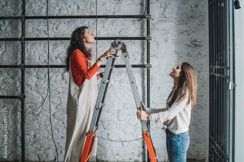 Two mid adult women on step ladders looking up in their new shop