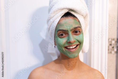 Woman with green face mask and head wrapped in towel standing in bathroom, smiling at camera.