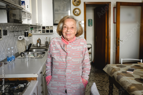 Old woman standing in her kitchen during Corona virus quarantine, smiling at camera. photo