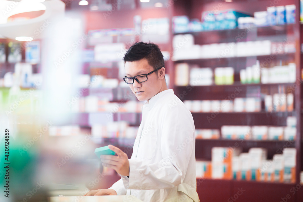 Male Asian pharmacist wearing glasses standing in front of shelves with medicines in a pharmacy.