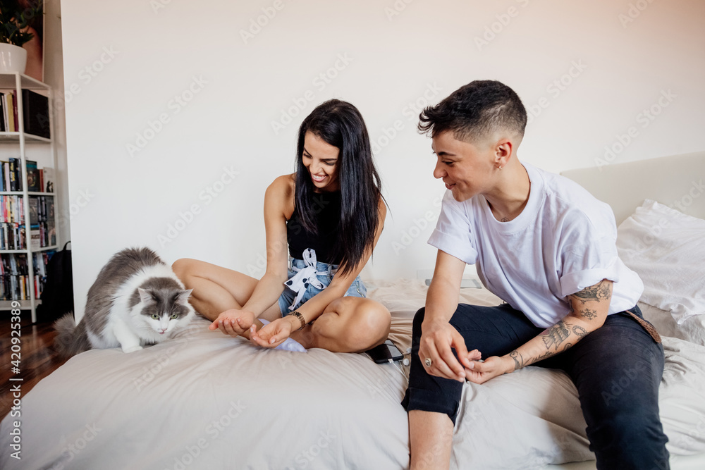 Young lesbian couple sitting on a bed, playing with cat. Young lesbian couple sitting on a bed, playing with cat.